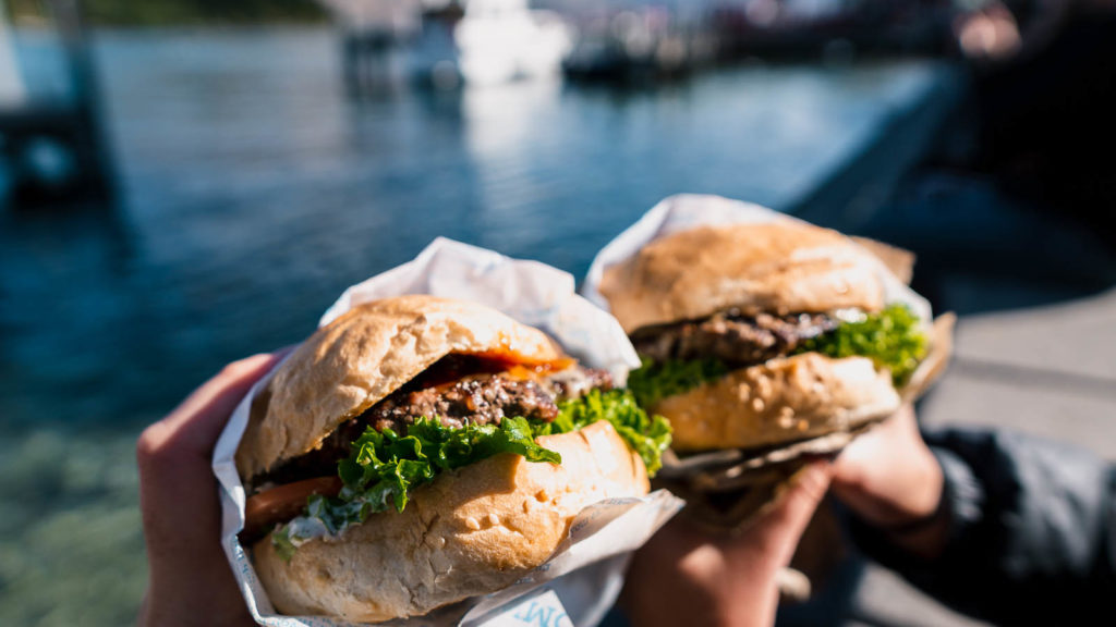 two hands holding burgers with water in the background