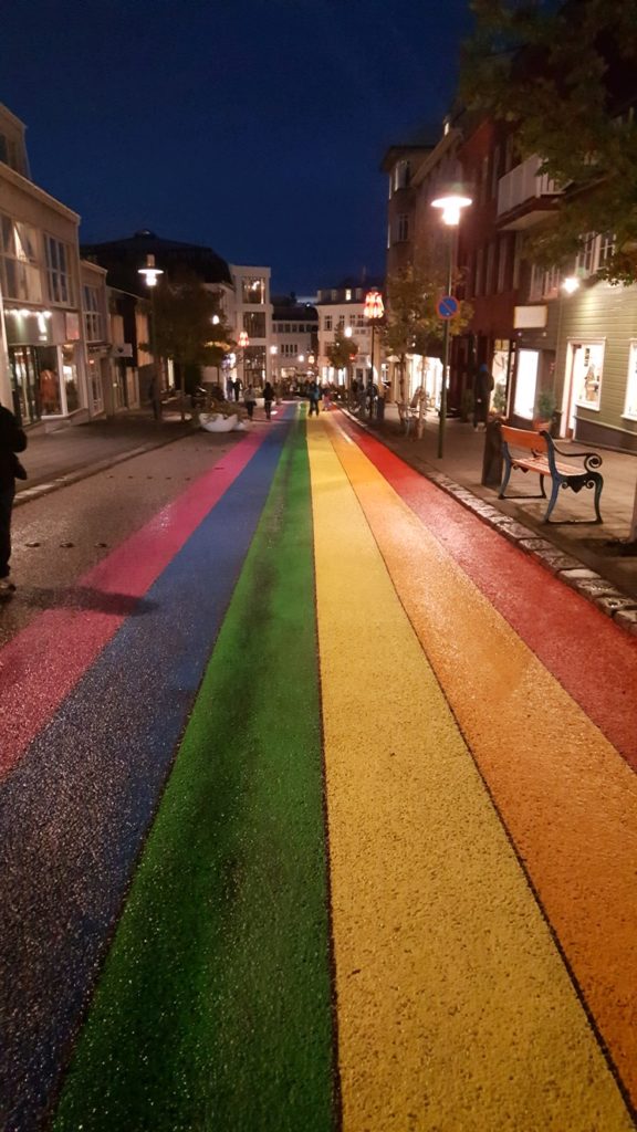 a rainbow colored street with buildings and people walking on it