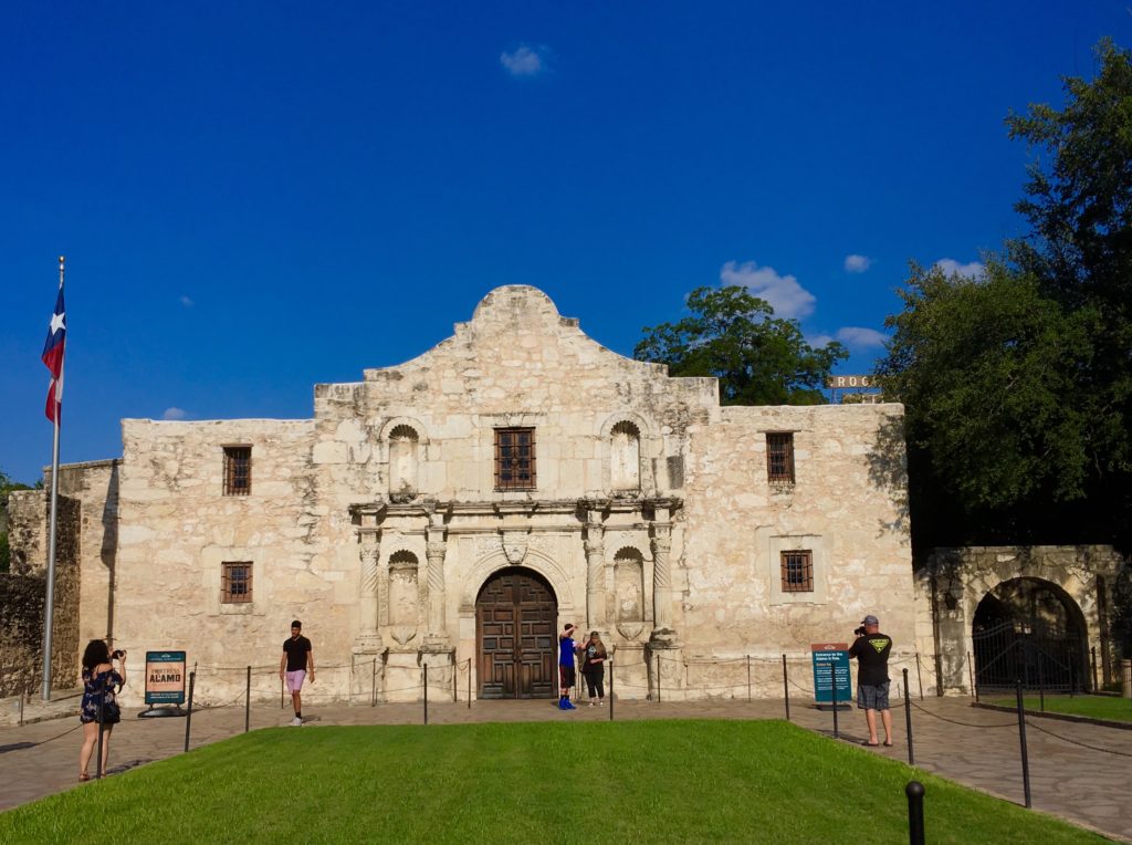 a stone building with a door and a stone wall with a stone fence and a green lawn with Alamo Mission in San Antonio in the background